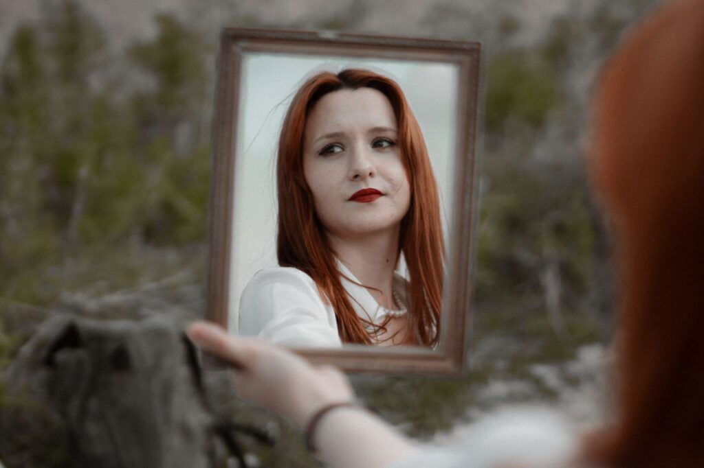 pexels photo 17733548 A young redhead woman holding a mirror outdoors, showcasing her reflection with red lips.