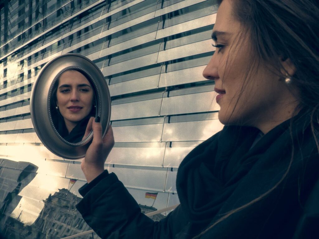 Главная Young woman in black jacket smiling at her reflection in a mirror outside a modern building.