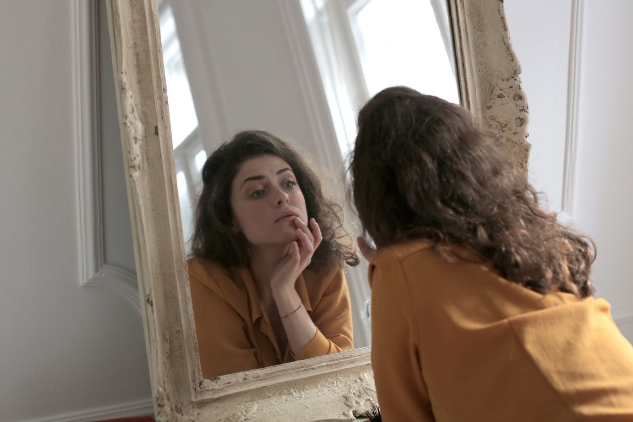 our-experience A woman in a mustard sweater thoughtfully gazing into a vintage mirror indoors.